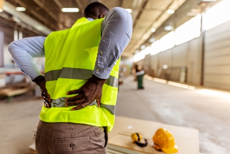 A worker wearing a safety vest holding their lower back inside a worksite. | Haight Stang, LLC